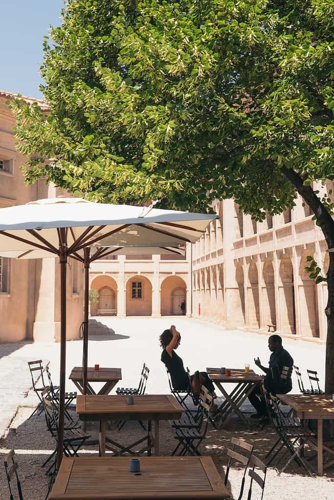 Two French beginners having a conversation in a courtyard. They're in the shade under a parasol and a large green tree.