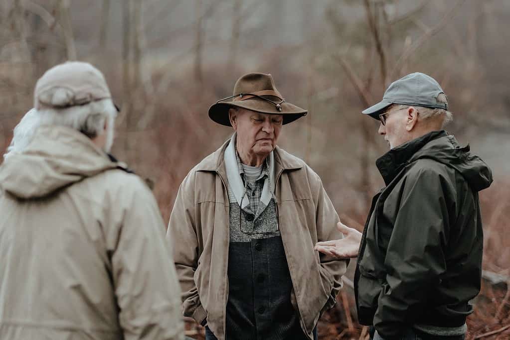 Trois hommes en tenue de campagne se tiennent et discutent en forêt, pratiquant leur 'anglais conversation' au milieu de la nature.