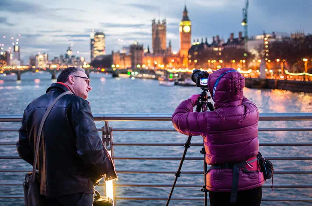 Un homme sur un pont à Londres engage une conversation en anglais avec une photographe en manteau violet, avec le Big Ben en arrière-plan - une illustration parfaite de l'utilisation du 'anglais guide de conversation' dans la vie réelle.